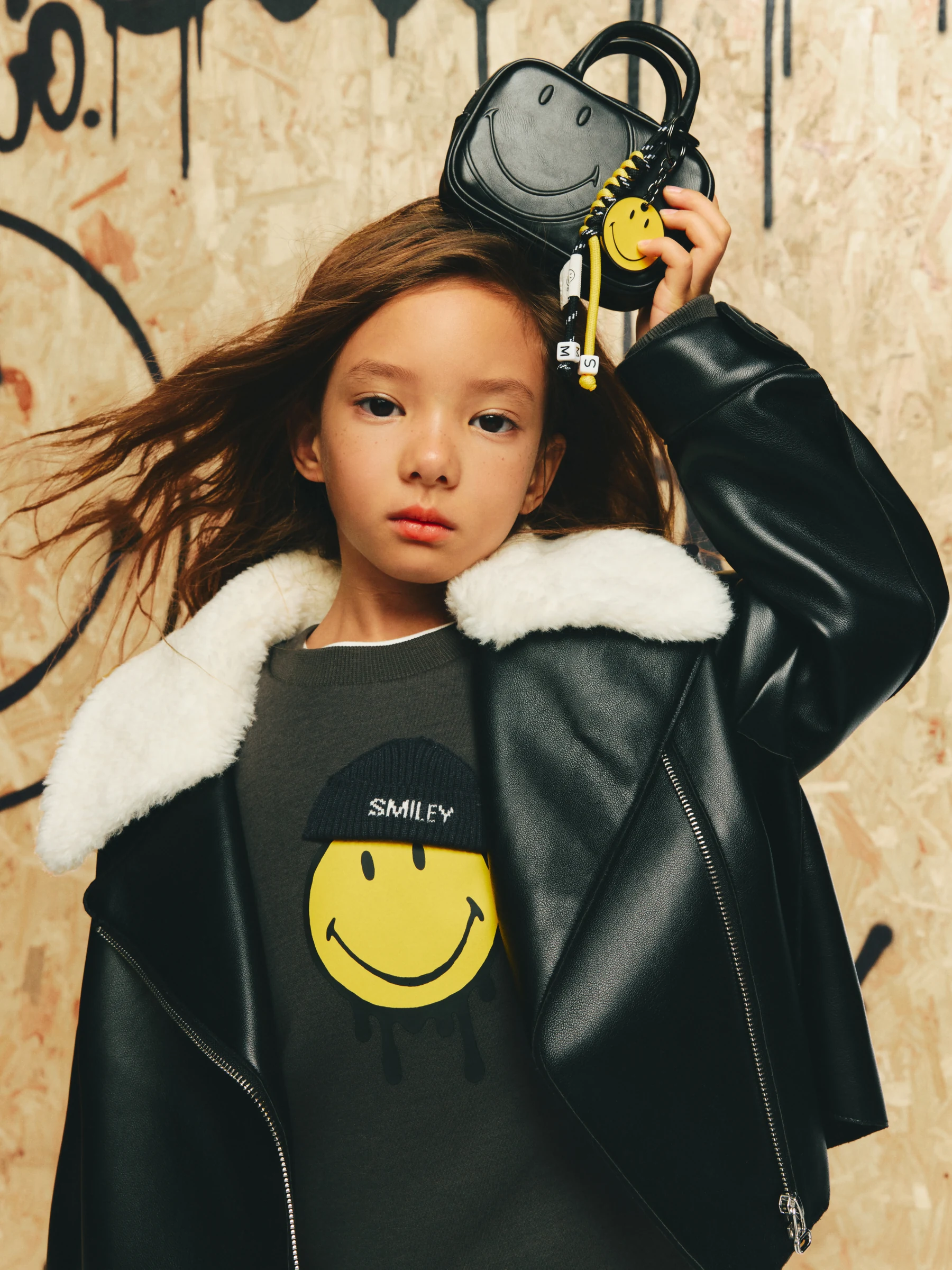 Girl in a shearling-trimmed jacket holding a smiley-detail bag